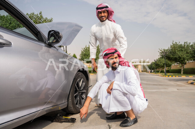 إصلاح بنشر العجلات، تعطل السيارة بشكل مفاجئ أثناء السير في الطريق، استخدام أدوات الصيانة اليدوية المناسبة، الفحص والتشييك على أجزاء المركبة, رجلان عربيان خليجيان سعوديان يرتديان الزي التقليدي يقفان في الكريق وبنظران الى شيء  ما، تثبيت الدولاب لوضع الصواميل