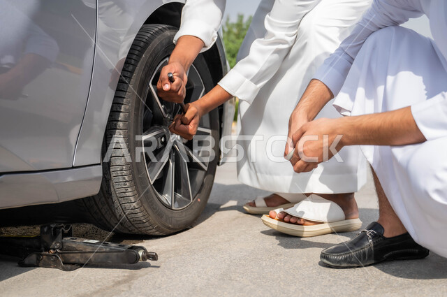 استخدام معدات الصيانة اليدوية المناسبة، تعطل المركبة بشكل مفاجئ أثناء السير في الطريق، تثبيت الدولاب لوضع الصواميل ،الفحص والتشييك على أجزاء السيارة, صورة مقربة لأيدي رجلان عربيان خليجيان سعوديان يرتديان الزي التقليدي السعودي يتساعدان في إصلاح بنشر العجلات