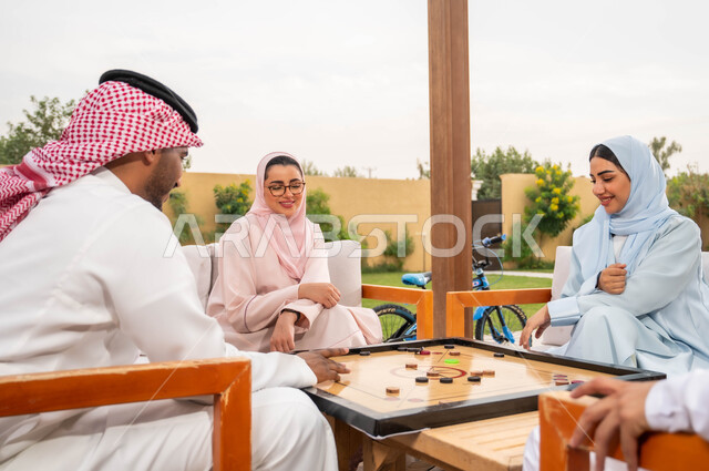 جمعات وأجواء عائلية سعيدة، مفهوم التحدي والتركيز، أنشطة ترفيهية وألعاب جماعية شعبية في المملكة، قضاء أوقات ممتعة برفقة الأهل والأصدقاء في إحدى المنتجعات السياحية، عائلة عربية خليجية سعودية تلعب بلعبة الكيرم (البليارد) المسلية في الهواء الطلق.