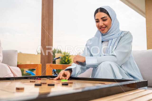 أجواء صيفية سعيدة، قضاء أوقات ممتعة في الهواء الطلق، التسلية والترفيه في إحدى الشاليهات السياحية، مفهوم التحدي والتركيز، صورة مقربة لامرأة عربية خليجية سعودية محجبة تلعب بلعبة الكيرم (البليارد) المسلية في الخارج بإيماءات السرور.