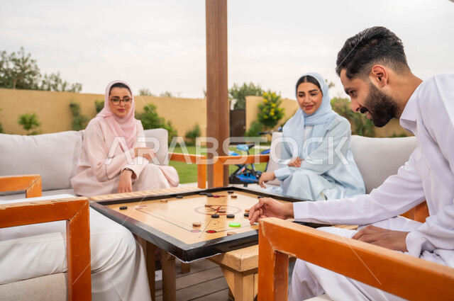 جمعات وأجواء عائلية سعيدة، مفهوم التحدي والتركيز، أنشطة ترفيهية وألعاب جماعية شعبية في المملكة، قضاء أوقات ممتعة برفقة الأهل والأصدقاء في إحدى المنتجعات السياحية، عائلة عربية خليجية سعودية تلعب بلعبة الكيرم (البليارد) المسلية في الهواء الطلق.