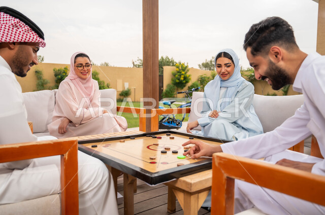 جمعات وأجواء عائلية سعيدة، مفهوم التحدي والتركيز، أنشطة ترفيهية وألعاب جماعية شعبية في المملكة، قضاء أوقات ممتعة برفقة الأهل والأصدقاء في إحدى المنتجعات السياحية، عائلة عربية خليجية سعودية تلعب بلعبة الكيرم (البليارد) المسلية في الهواء الطلق.