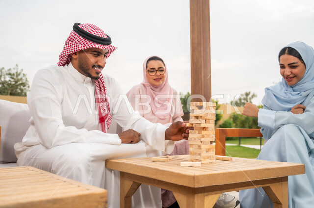 إيماءات المتعة والسعادة في إحدى الشاليهات في العطلة الصيفية، ممارسة الأنشطة والألعاب الجماعية المسلية لتنمية مهارات التفكير والذكاء، عائلة عربية خليجية سعودية تلعب لعبة جينغا في الهواء الطلق، رص القطع والألواح الخشبية