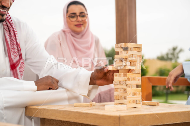 رص القطع والألواح الخشبية، إيماءات المتعة والسعادة في إحدى الشاليهات في العطلة الصيفية، ممارسة الأنشطة والألعاب الجماعية المسلية لتنمية مهارات التفكير والذكاء، عائلة عربية خليجية سعودية تلعب لعبة جينغا في الهواء الطلق