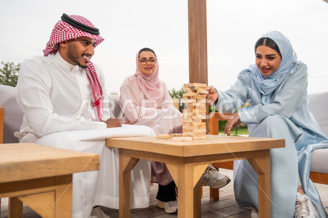 إيماءات المتعة والسعادة في إحدى الشاليهات في العطلة الصيفية، ممارسة الأنشطة والألعاب الجماعية المسلية لتنمية مهارات التفكير والذكاء، عائلة عربية خليجية سعودية تلعب لعبة جينغا في الهواء الطلق، رص القطع والألواح الخشبية