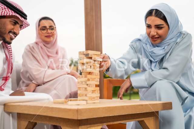 رص القطع والألواح الخشبية، إيماءات المتعة والسعادة في إحدى الشاليهات في العطلة الصيفية، ممارسة الأنشطة والألعاب الجماعية المسلية لتنمية مهارات التفكير والذكاء، عائلة عربية خليجية سعودية تلعب لعبة جينغا في الهواء الطلق