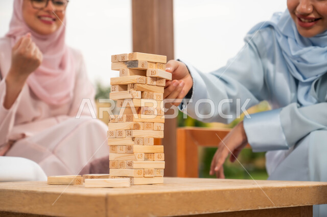 ممارسة الأنشطة والألعاب المسلية مع الأصدقاء في إحدى الشاليهات في العطلة الصيفية، صورة مقربة لسيدتان عربيتان خليجيتان سعوديتان تلعبان لعبة جينغا في الهواء الطلق بإيماءات المتعة والسعادة والحماس، رص القطع والألواح الخشبية