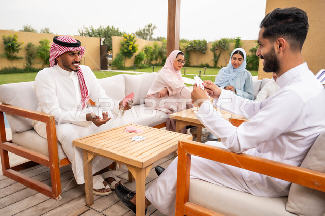 مفهوم التركيز والتحدي في لعب الشدة، قضاء وقت ممتع في ممارسة الأنشطة والألعاب المسلية، الاستمتاع والتسلية مع الأهل والأصدقاء في إحدى الشاليهات في العطلة الصيفية، رجلان عربيان خليجيان سعوديان يلعبان لعبة الورق بإيماءات السعادة والسرور