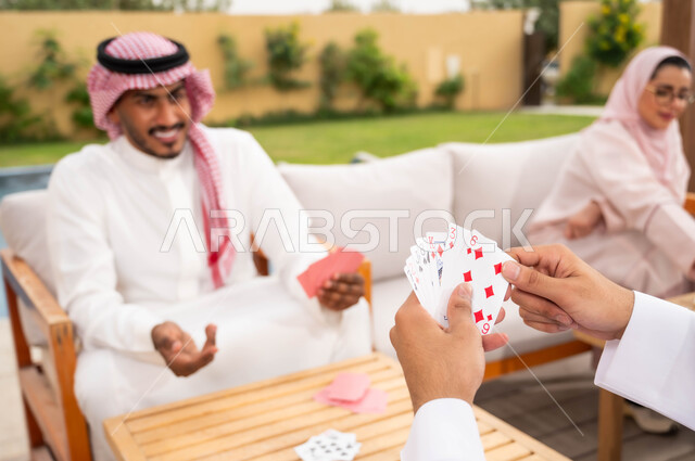 ممارسة الأنشطة والألعاب الجماعية المسلية، التسلية والاستمتاع مع الأهل والأصدقاء في إحدى الشاليهات في العطلة الصيفية، مفهوم التركيز والتحدي في لعب الشدة، رجلان عربيان خليجيان سعوديان يلعبان لعبة الورق بإيماءات السعادة والسرور