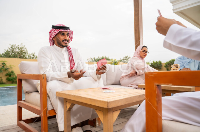 التسلية والاستمتاع مع الأهل والأصدقاء في إحدى الشاليهات في العطلة الصيفية، مفهوم التركيز والتحدي في لعب الشدة، قضاء وقت ممتع في ممارسة الأنشطة والألعاب المسلية، رجلان عربيان خليجيان سعوديان يلعبان لعبة الورق بإيماءات السعادة والسرور
