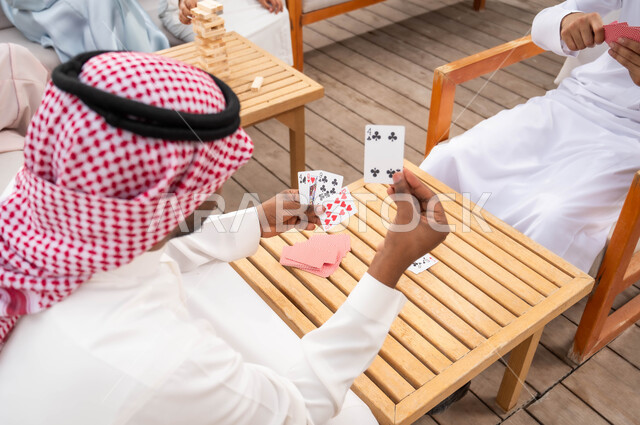 التسلية والاستمتاع مع الأهل والأصدقاء في إحدى الشاليهات في العطلة الصيفية، مفهوم التركيز والتحدي في لعب الشدة، قضاء وقت ممتع في ممارسة الأنشطة والألعاب المسلية، رجلان عربيان خليجيان سعوديان يلعبان لعبة الورق بإيماءات السعادة والسرور