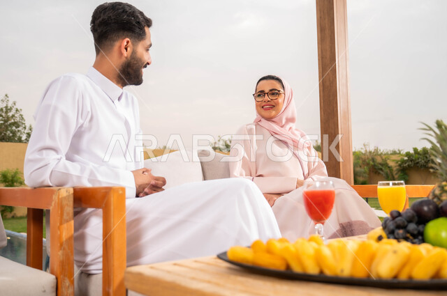 مفهوم العلاقات الزوجية السعيده، أطباق الفواكه المتنوعه والعصائر الطبيعيه المنعشة على الطاولة، الاستمتاع في أوقات الفراغ في إحدى الشاليهات، التنزه في الهواء الطلق، زوجان عربيان خليجيان سعوديان يقومان بتبادل الأحاديث والدردشات