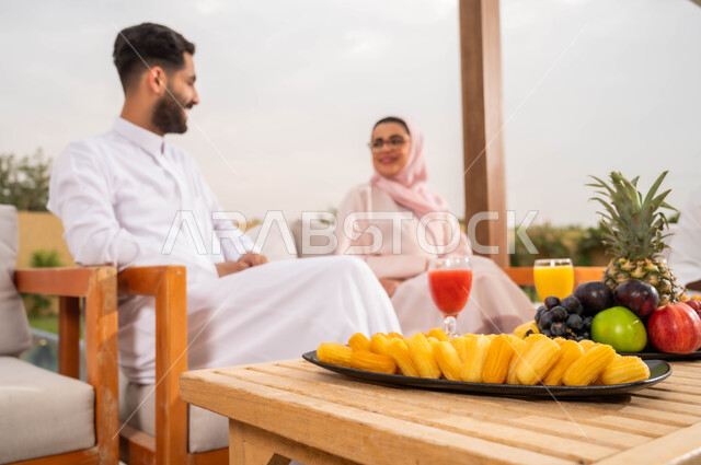 مفهوم العلاقات الزوجية السعيده، أطباق الفواكه المتنوعه والعصائر الطبيعيه المنعشة على الطاولة، الاستمتاع في أوقات الفراغ في إحدى الشاليهات، التنزه في الهواء الطلق، زوجان عربيان خليجيان سعوديان يقومان بتبادل الأحاديث والدردشات