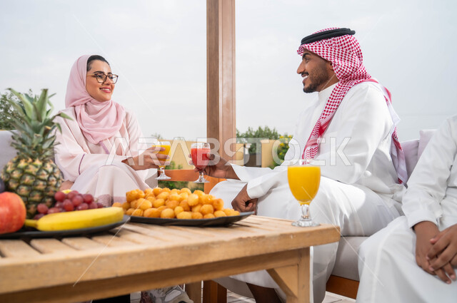 أطباق الفواكه المتنوعه والعصائر الطبيعيه المنعشة على الطاولة، الاستمتاع في إحدى الشاليهات، التنزه في الهواء الطلق، زوجان عربيان خليجيان سعوديان يقومان بتبادل الأحاديث والدردشات