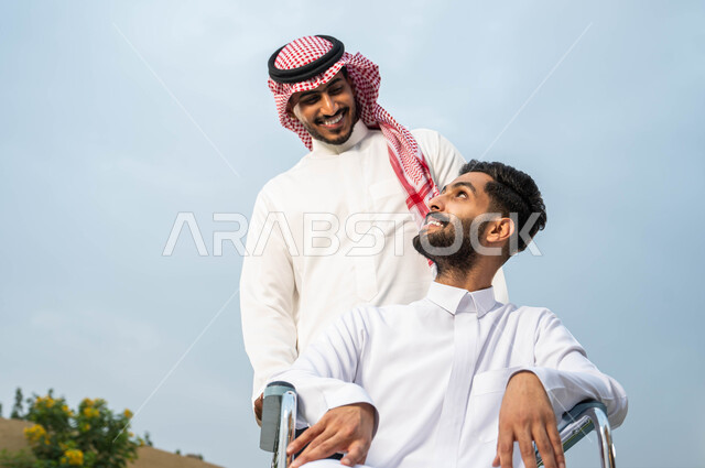 نمط حياة أصحاب الهمم في المملكة، التنزه في الهواء الطلق، مساعدة ذوي الإعاقة الحركية على التنقل بسهولة, رجل عربي خليجي سعودي يرتدي الزي التقليدي برفقة صديقه في إحدى الحدائق، صورة مقربة لشاب مقعد يجلس على كرسي متحرك بإيماءات السرور، مفهوم رعاية المرضى والمعاقين