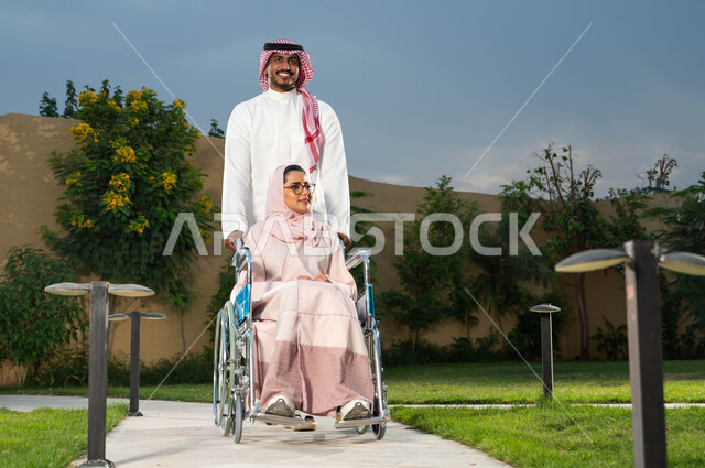 التنزه في الهواء الطلق، مساعدة مرضى ذوي الإعاقة الحركية على التنقل بسهولة, رجل عربي خليجي سعودي يرتدي الزي التقليدي برفقة زوجته في إحدى الحدائق، امرأة سعودية مقعدة تجلس على كرسي متحرك، نمط الحياة في المملكة