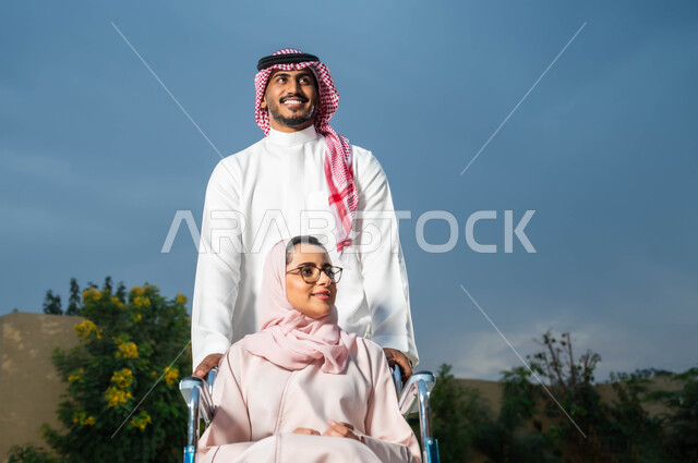 نمط حياة أصحاب الهمم في المملكة، التنزه في الهواء الطلق، مساعدة مرضى ذوي الإعاقة الحركية على التنقل بسهولة, رجل عربي خليجي سعودي يرتدي الزي التقليدي برفقة زوجته في إحدى الحدائق، امرأة سعودية مقعدة تجلس على كرسي متحرك