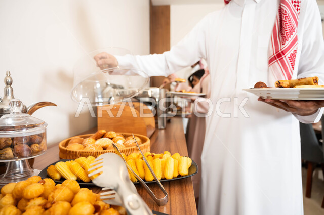 اطباق متنوعه من المأكولات والمشروبات، مفهوم الخدمة الذاتية في المطاعم السعودية، صورة مقربة لرجل عربي خليجي سعودي يرتدي الزي التقليدي يقوم بسكب الطعام من البوفيه في المطعم, حلويات شعبية طازجة
