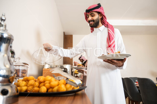حلويات شعبية طازجة، اطباق متنوعه من المأكولات والمشروبات، مفهوم الخدمة الذاتية في المطاعم السعودية، صورة مقربة لرجل عربي خليجي سعودي مبتسم يرتدي الزي التقليدي يقوم بسكب الطعام من البوفيه في المطعم بإيماءات السرور