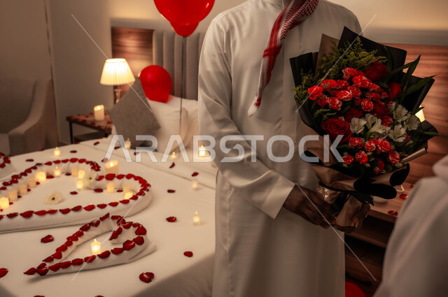 سرير مزين بالشموع والورود الحمراء للاحتفال بيوم الفلنتاين، الاهتمام بالمناسبات الخاصة، الاحتفال بذكرى الزواج، زوج عربي خليجي سعودي يقوم بتقديم باقة من الزهور لزوجته، تزيين غرفة النوم بطريقة رومانسية.