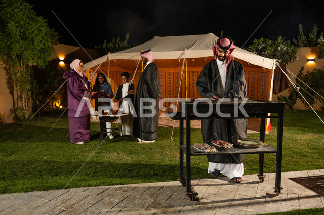 التجمع حول الموقد للتدفئة، التسامر وتبادل الأحاديث في كشتة بر وتخييم في الأجواء الشتوية، عائلة عربية خليجية سعودية يقيمون حفلة شواء في رحلة برية في إحدى الشاليهات السياحية في المملكة، قضاء أوقات عائلية ممتعة