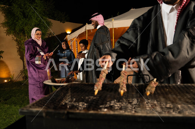 التسامر وتبادل الأحاديث في كشتة بر وتخييم في الأجواء الشتوية، عائلة عربية خليجية سعودية يقيمون حفلة شواء في رحلة برية في إحدى الشاليهات السياحية في المملكة، قضاء أوقات عائلية ممتعة، التجمع حول الموقد للتدفئة