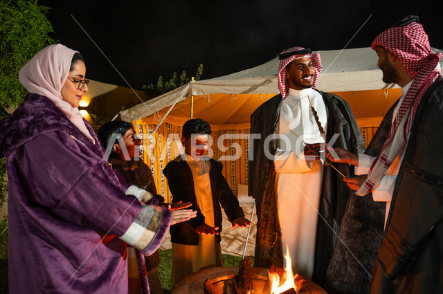 التجمع حول الموقد للتدفئة، التسامر وتبادل الأحاديث في كشتة بر وتخييم في الأجواء الشتوية، عائلة عربية خليجية سعودية يقيمون حفلة شواء في رحلة برية في إحدى الشاليهات السياحية في المملكة، قضاء أوقات عائلية ممتعة
