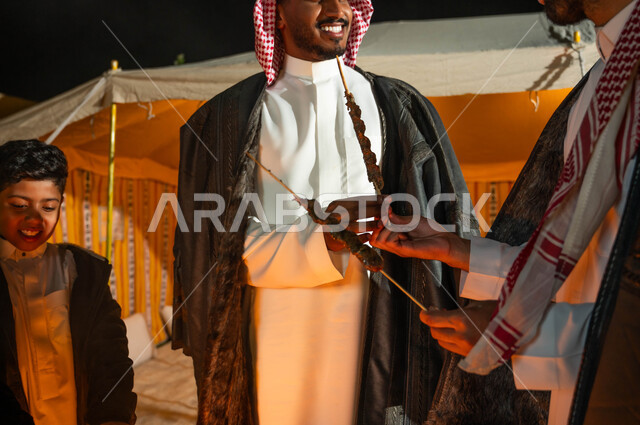 تبادل الأحاديث والتسامر في كشتة بر وتخييم في الأجواء الشتوية، التجمع حول الموقد للتدفئة، صورة مقربة لرجلين عربيين خليجيين سعوديين يقيمان حفلة شواء في رحلة برية في إحدى الشاليهات السياحية في المملكة، قضاء أوقات عائلية ممتعة