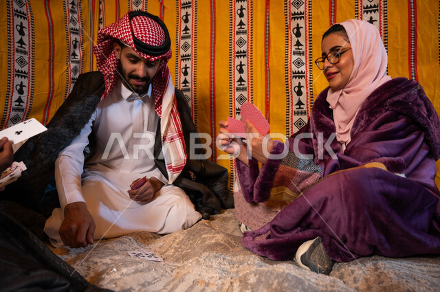 مفهوم التركيز والتحدي في لعبة الكوتشينه، التسلية والترفيه والاستمتاع باللعب الجماعي, عائلة عربية خليجية سعودية يلعبون لعبة الورق, قضاء أوقات ممتعه في كشتة برية شتوية في صحاري السعودية.