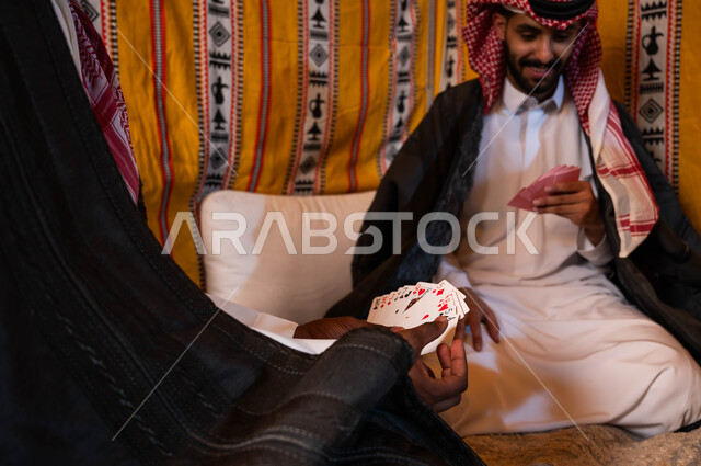 قضاء أوقات ممتعه في كشتة برية شتوية في صحاري المملكة، التسلية والترفيه والاستمتاع باللعب الجماعي, مفهوم التركيز والتحدي في لعبة الكوتشينه، صورة مقربة لرجلين عربيين خليجيين سعوديين يرتديان الزي التقليدي يلعبان لعبة الورق.