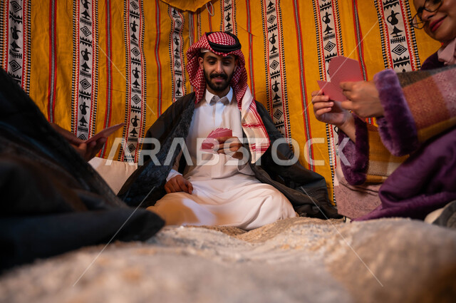 التسلية والترفيه والاستمتاع باللعب الجماعي, مفهوم التركيز والتحدي في لعبة الكوتشينه، صورة مقربة لعائلة عربية خليجية سعودية يلعبون لعبة الورق, قضاء أوقات ممتعه في كشتة برية شتوية في صحاري المملكة.