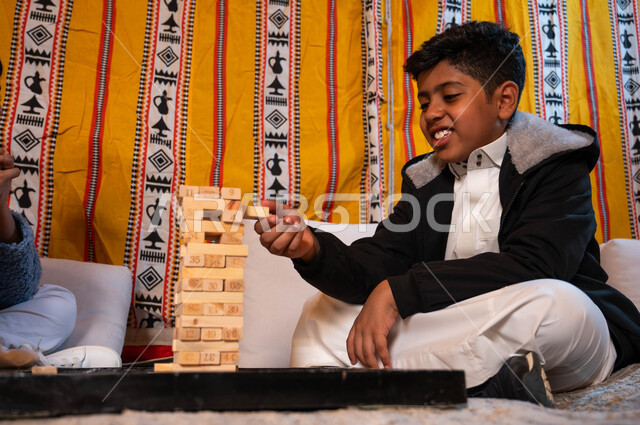 جلسة بر في الأجواء الشتوية، ألعاب عائلية مسلية، قضاء إجازة ممتعة في اللعب والتسلية، ولد عربي خليجي سعودي لعب لعبة جينغا، رص الألواح الخشبية، أنشطة ترفيهية لتنمية مهارات التفكير والذكاء عند الأطفال.