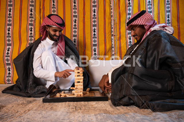 ألعاب جماعية مسلية، قضاء إجازة ممتعة في كشتة برية شتوية في صحاري السعودية، شابان عربيان خليجيان سعوديان يلعبان لعبة جينغا بإيماءات الفرح والسرور، رص الألواح الخشبية، أنشطة ترفيهية لتنمية مهارات التفكير والذكاء.
