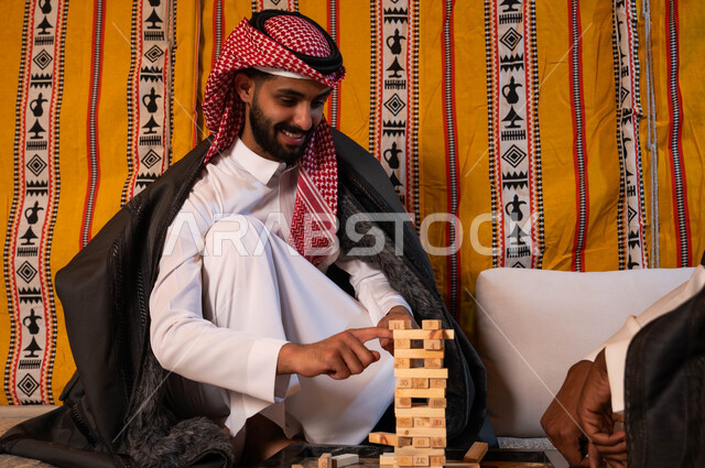 Spending an enjoyable vacation in a wild winter country in the deserts of Saudi Arabia, entertaining group games, two young Saudi Gulf Arabs playing the Jenga game with gestures of joy and pleasure, stacking wooden boards, recreational activities to develop thinking skills and intelligence.