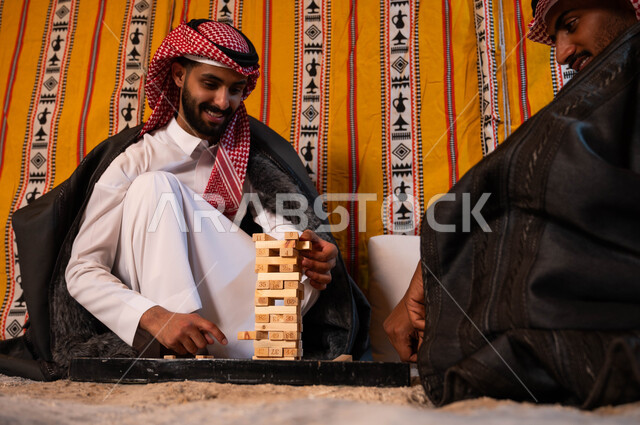 ألعاب جماعية مسلية، قضاء إجازة ممتعة في كشتة برية شتوية في صحاري السعودية، شابان عربيان خليجيان سعوديان يلعبان لعبة جينغا بإيماءات الفرح والسرور، رص الألواح الخشبية، أنشطة ترفيهية لتنمية مهارات التفكير والذكاء.