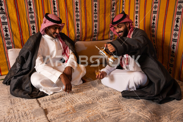 مفهوم حسن الضيافه والاستقبال، صب القهوة من الدلة النحاسية، إيماءات البشاشة والترحيب، التسامر وتبادل الحديث في كشتة شعبية شبابية، صديقان عربيان خليجيان سعوديان في جلسة بر وتخييم خارجية في الأجواء الشتوية