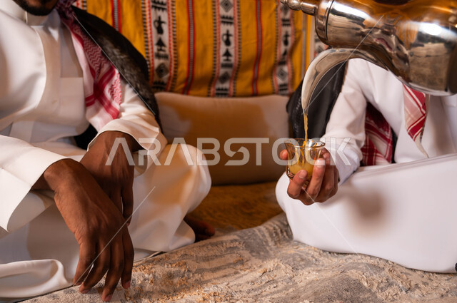 صب القهوة من الدلة النحاسية، إيماءات البشاشة والترحيب، مفهوم حسن الضيافه والاستقبال، التسامر وتبادل الحديث في كشتة شعبية شبابية، صورة مقربة لصديقين عربيين خليجيين سعوديين في جلسة بر وتخييم خارجية في الأجواء الشتوية
