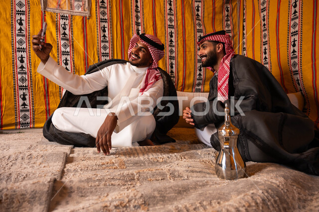 توثيق اللحظات السعيدة، مفهوم حسن الضيافه والاستقبال، رجل عربي خليجي سعودي يقوم بالتقاط صور السيلفي التذكارية مع صديقه باستخدام الهاتف الجوال، كشتة شعبية شبابية، صديقان عربيان خليجيان سعوديان في جلسة بر وتخييم خارجية في الأجواء الشتوية
