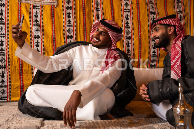كشتة شعبية شبابية، توثيق اللحظات السعيدة، مفهوم حسن الضيافه والاستقبال، رجل عربي خليجي سعودي يقوم بالتقاط صور السيلفي التذكارية مع صديقه باستخدام الهاتف الجوال، صديقان عربيان خليجيان سعوديان في جلسة بر وتخييم خارجية في الأجواء الشتوية