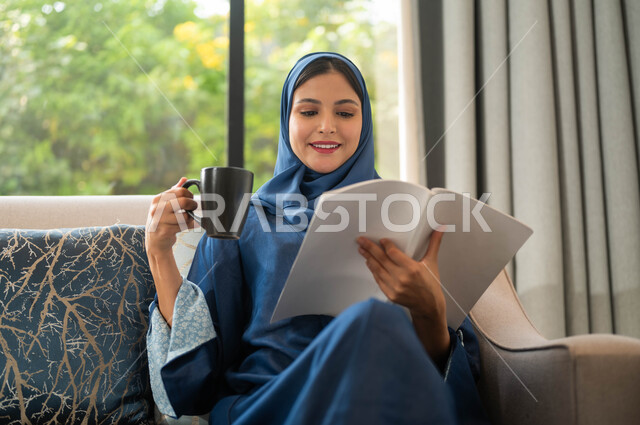 ايماءات التركيز والاستمتاع، تمضية أوقات الفراغ بالتعلم والتثقيف الذاتي، صورة مقربة لسيدة عربية سعودية خليجية ترتدي العباءة الملونة تحمل بيدها كتاب وتحتسي كوب من القهوة, قضاء أوقات هادئة في المنزل, قراءة الروايات والقصص