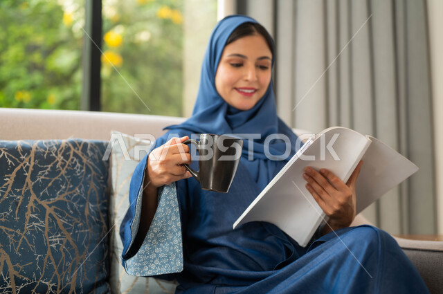 تمضية أوقات الفراغ بالتعلم والتثقيف الذاتي، صورة مقربة لامراة عربية سعودية خليجية ترتدي العباءة الملونة تحمل بيدها كتاب وتحتسي كوب من القهوة, قضاء أوقات هادئة في المنزل, قراءة الروايات والقصص, ايماءات التركيز والاستمتاع