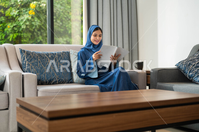 قراءة القصص والروايات الشيقة والممتعة، امرأة عربية خليجية سعودية تجلس في غرفة المعيشة وتحمل بيدها كتاب، الاسترخاء والاستمتاع بالأجواء الهادئة، اكتساب الثقافة والمعرفة الفكرية