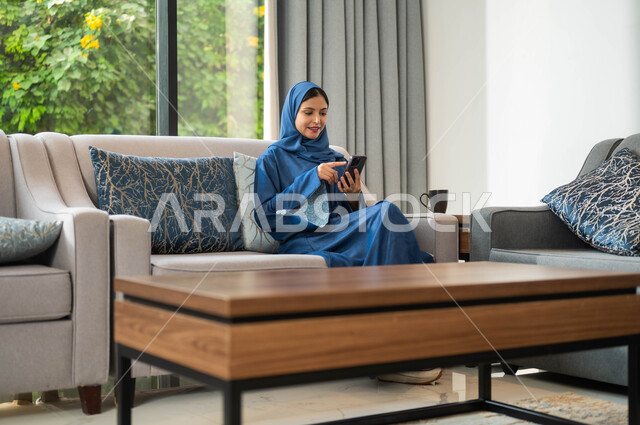 استخدام التكنولوجيا المتطورة والحديثة، سيدة عربية خليجية سعودية محجبة مبتسمة تجلس في المنزل و تمسك بيدها الهاتف المحمول، تصفح تطبيقات وبرامج التواصل الاجتماعي، قضاء أوقات مسلية وممتعة