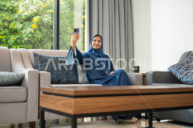 توثيق اللحظات السعيدة، إجراء مكالمة فيديو عبر الجوال, دردشات وحوارات ودية مع الأصحاب، امراة عربية خليجية سعودية محجبة تلتقط صورة سيلفي تذكارية بالهاتف المحمول، مفهوم صلة الرحم و التواصل مع الأصدقاء والأقارب
