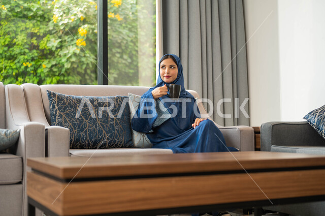 غرفة معيشة بتصاميم عصرية جذابة و ديكورات فاخرة، ايماءات الهدوء والاستمتاع بالمشروبات الدافئة داخل المنزل، امراة عربية سعودية خليجية تجلس على الأريكة وتمسك بيدها كوب من القهوة
