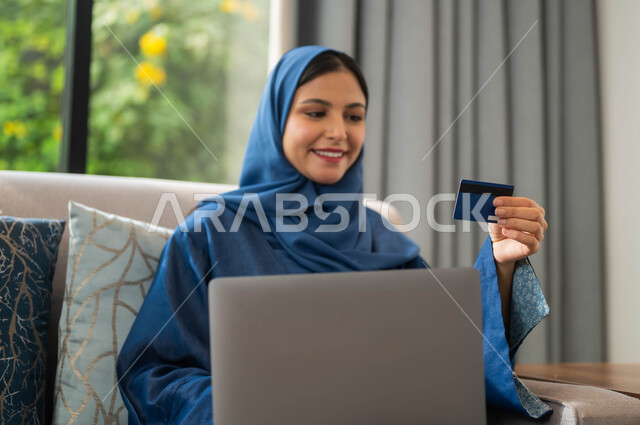 صورة مقربة لإمرأة عربية سعودية خليجية محجبة بالزي التقليدي مبتسمة تجلس على الأريكة تعمل على جهاز اللابتوب تنظر بإيماءات السعادة والسرور، التطور والتكنولوجيا الحديثة، مفهوم الدفع الالكتروني عبر تطبيقات الحاسب المحمول، استخدام البطاقة البنكية الالكترونية