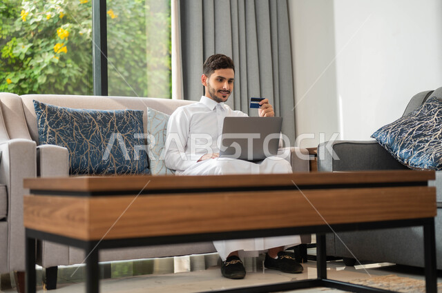 Following up on banking services through the laptop, paying through the credit card, purchasing through electronic applications, a young Gulf Arab Saudi man holding the bank card in his hand and sitting on the sofa in the living room.
