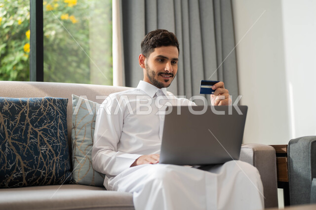 استخدام الأجهزة الالكترونية والتقنيات الحديثة، رجل عربي خليجي سعودي مبتسم يرتدي الثوب التقليدي يمسك بيده البطاقة البنكية، انجاز المهام عبر الانترنت، إدارة الاعمال والحسابات عن بعد