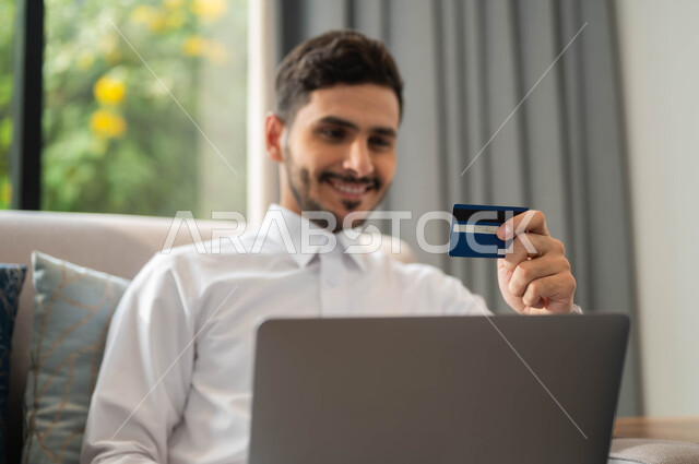 الدفع عبر تطبيقات اللابتوب، استخدام البطاقة البنكية الالكترونية، صورة مقربة لرجل عربي سعودي خليجي مبتسم الثوب التقليدي يجلس على كنبة مريحة يعمل على جهاز الحاسوب المحمول، إمكانية تسديد الفواتير عبر الانترنت
