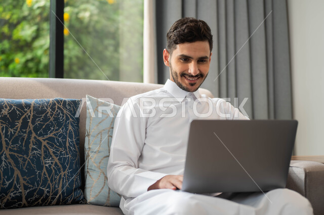 مفهوم العمل عن بعد، انجاز الأعمال من المنزل عبر الانترنت، تصفح برامج وتطبيقات الابتوب، بورتريه  لرجل عربي سعودي خليجي مبتسم يرتدي الثوب التقليدي يجلس على أريكة مريحة يقوم باستخدام الحاسوب المحمول، الأجهزة والتقنيات المتطورة